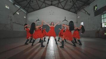 A group of six dancers in red dresses performing in an industrial space with high ceilings and wooden beams. The setting is minimalistic, with natural light illuminating the scene. video
