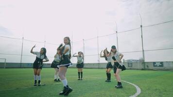 A group of five young women performing a dance routine on a grassy field, wearing stylish outfits. The background features a sports field with a cloudy sky. video
