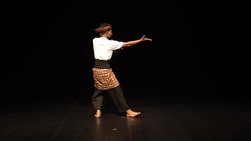 A male dancer performing on stage, wearing traditional attire with a checkered sarong and a white shirt. The stage is dimly lit, highlighting the dancer's movements. video