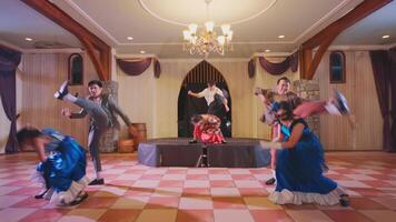 A lively dance performance featuring five dancers on stage in a decorated venue. The dancers wear colorful costumes, showcasing a mix of styles. The atmosphere is festive with a chandelier overhead. video
