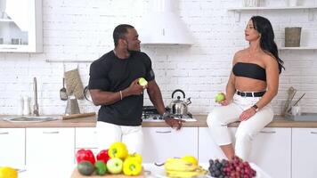 Sporty couple having a conversation at home. Caucasian woman sitting on the kitchen table and talking to afro man. Multi ethnic couple eating fresh apples. Healthy eating. video