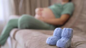Beautiful tiny knitted socks for future child. Baby socks on blurred background of a living-room with a pregnant woman sitting on a couch and stroking her stomach. video