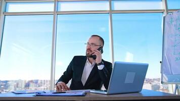 Portrait of entrepreneur at workplace. Confident businessman in glasses talking the phone while sitting in front of a laptop in office. video