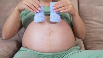 Beautiful blue knitted socks in hands of a pregnant woman. Big belly of a future mother laying on a sofa at home. Close-up. Happy pregnancy. video