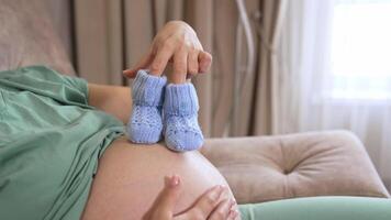 Pregnant woman playing with tiny baby socks. Future mom with blue knitted socks stepping on her belly while relaxing at home. Expectant mother enjoys pregnancy time. video