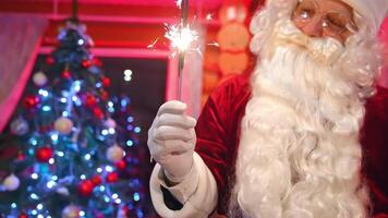 Portrait of Santa Claus with sparkler lights. Happy Santa with white beard wearing glasses standing on Christmas tree backdrop and looking on camera. video