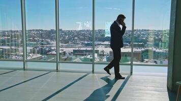 Young businessman talking the phone in office. Handsome entrepreneur in suit walking near large office windows and having a phone conversation. video