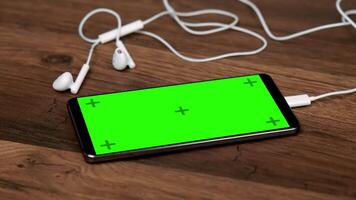 Modern smartphone with green screen mockup and white earphones on natural wooden surface in minimalist setup video