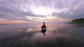 FPV drone shot of woman riding motorcycle on wide black sand beach in Bali at sunset in slow motion. Peaceful, cinematic scene symbolizing freedom, exploration, and slow travel lifestyle. video