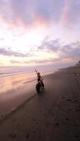 FPV drone shot of woman riding motorcycle on wide black sand beach in Bali at sunset in slow motion. Peaceful, cinematic scene symbolizing freedom, exploration, and slow travel lifestyle. video