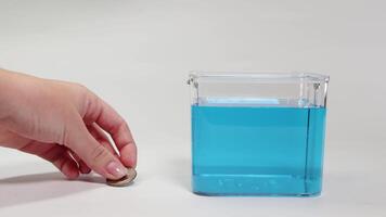 A hand drops a metal coin into a clear container filled with blue liquid, demonstrating buoyancy and density video