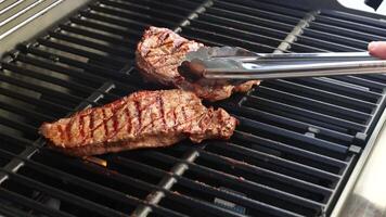 Two beef steaks being grilled on a barbecue, with tongs used to flip them. Bright outdoor lighting enhances the scene video