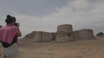 Tourist capturing photo of historic stone fortress walls on dry grassy field under cloudy sky. video