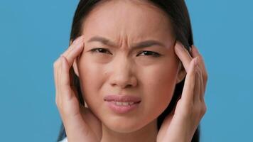 A millennial Asian woman expresses discomfort while touching her temples. The backdrop is a vibrant blue, emphasizing her facial expression and isolated presence. video
