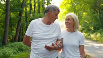 Elderly couple in their 60s walking through a vibrant green park. Surrounded by trees, they share smiles while embracing an active lifestyle amid nature. video
