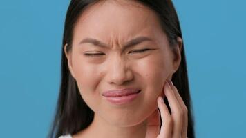 Toothache. A millennial woman of Asian descent grimaces as she touches her cheek, showing signs of discomfort. She is isolated against a solid blue background, focusing on her expression. video