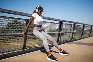 Outdoor shot of muscular African-American woman doing reverse push ups exercises with headphones, enjoying music while staying active. photo