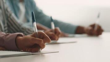 Diverse group of college students focus on their studies, writing notes with pens on notebooks. They are collaborating in a bright learning environment, emphasizing education and teamwork. video