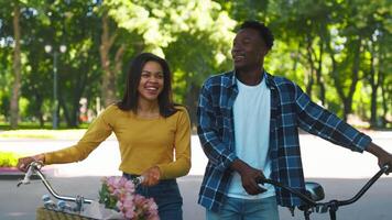 A young African American couple rides bicycles through a vibrant park filled with greenery, sharing laughter and joy. The woman holds a bouquet of flowers, enhancing the cheerful atmosphere. video