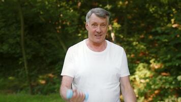An elderly man is exercising in a green park, embracing an active lifestyle. Grandfather enjoying the fresh air and beautiful surroundings during outdoor exercise routine with dumbbells video
