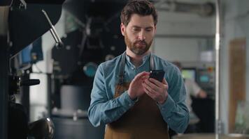 Caucasian man worker with smartphone in coffee roasted fabric industry working stand near roasting machine using mobile phone male small business owner inspecting quality browsing cellphone chatting video