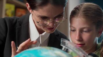 Close up learning together Caucasian woman female professor tutor and little girl student school child kid learner pupil schoolgirl talking studying geography looking at globe world map planet indoor video