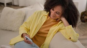 Ethnic African American woman smiling relaxed girl on home sofa couch using mobile phone shopping online purchase scrolling social media gadget addict female shopper browsing internet texting messages video