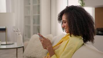 Beautiful African American woman relaxing on cozy couch use mobile phone application at home living room happy smiling ethnic curly girl chatting in social network addict internet playing online game video