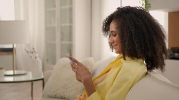 Smiling African American girl woman play mobile game on home couch in living room happy relaxed ethnic female teenager scrolling smartphone social networks addict internet dating apps side view video