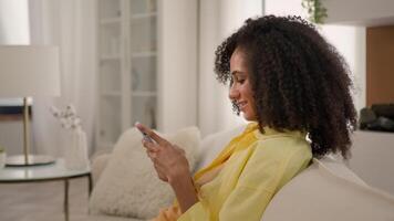 African American ethnic woman smiling 20s gen z girl on home couch relax scrolling smartphone buying online store internet order browsing mobile apps phone gadget addiction social media in apartment video