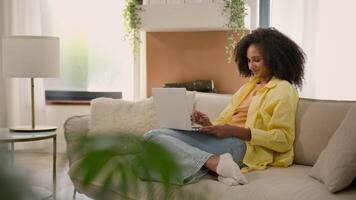 African American woman homeowner relaxing on couch with laptop device online chatting web surfing internet studying with computer at home ethnic girl businesswoman freelance business work e-learning video