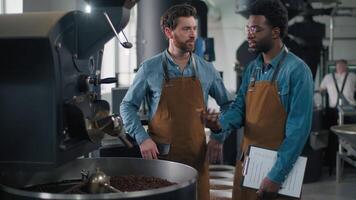 Professional multinational two Caucasian African men workers in uniform apron conversation coffee roasting machinery talking factory males inspection work production workspace process communication video
