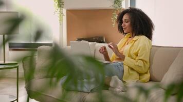 African American ethnic woman female home owner talk with laptop conference app online distant call business chat pleasant communication smiling girl on couch using computer in house with plants video