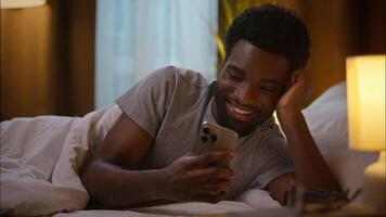 Happy resting African American ethnic man smiling male cheerful guy user client lying bed blanket using mobile phone smartphone browsing internet app relaxing watch funny at home bedroom night video