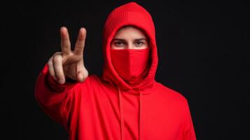 Young man in a red hoodie and mask showing peace sign against a dark background photo