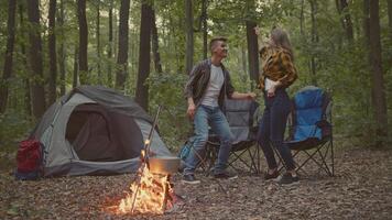 A couple enjoys a joyful moment dancing near a campfire in a lush forest. Their tent and camping chairs are set up, showcasing an active lifestyle amid nature's beauty. video