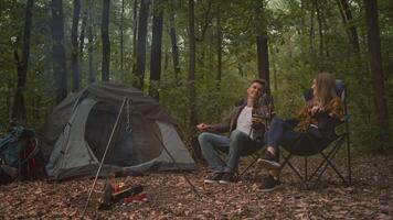 Two friends relax around a campfire in a lush forest, sharing stories and laughter. Their tent stands nearby, showcasing a perfect outdoor experience amidst nature. video