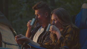 In the tranquil forest, a couple relaxes in camping chairs, sharing a peaceful moment by the campfire while sipping from metal mugs, checking road map video