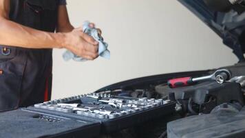Auto mechanic cleaning dirty hands with cloth after completing car engine repair, standing near open car hood. Man finishing work in auto repair shop. Vehicle maintenance concept video