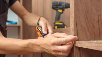 Close-up of man measuring wooden wardrobe with tape measure during furniture assembly, with tools and screws on shelf in background. Concept of home renovation and DIY furniture project video