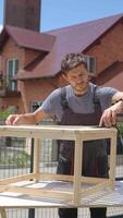 Man assembling wooden furniture frame outdoors on sunny day, wearing overalls. Carpenter assembling DIY terrace table in backyard. Handmade furniture making video