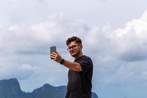 Young man taking selfie outdoors against scenic backdrop of mountains and clouds, showcasing moment of joy and connection with nature and technology. Man taking selfie against sky photo