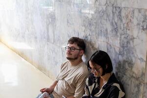 Two tired young adults in glasses are sitting against stylish marble wall. Tired tourists man and woman on tour sitting on floor and resting. Exhaustion and fatigue while traveling photo