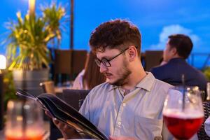 Young man with glasses is focused on reading menu at rooftop restaurant, surrounded by soft lighting and evening ambiance, with drinks and greenery enhancing atmosphere. Restaurant terrace at sunset photo