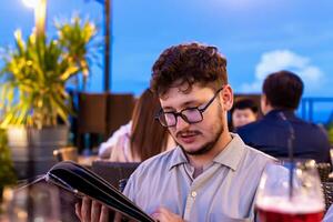 Young man with glasses is focused on reading menu in lively outdoor restaurant, surrounded by greenery and softly lit ambiance, creating relaxed dining atmosphere. Restaurant terrace at sunset photo