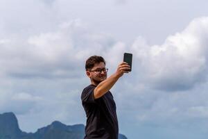 Young man with glasses is taking selfie outdoors against scenic backdrop of mountains and clouds, capturing moment of joy and adventure in nature. Male individual is taking selfie in outdoor setting photo