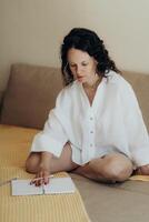 Woman in white linen shirt sits cross legged on beige couch journaling in notebook with pastel pencils in soft light expressing calm and introspective quiet moment of mindful reflection photo