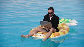 Young man sitting on an inflatable in a pool using a laptop. Concept remote work, freelance lifestyle and summer freedom. video
