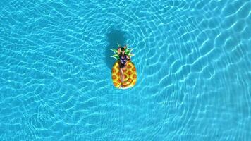 Concept summer fun, relaxation, tropical vibes and leisure. Top view of woman on a big pineapple float in a clear blue pool, shot from drone. video
