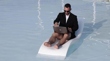 homem dentro roupa de banho e uma blazer usando uma computador portátil enquanto reclinável de a piscina. conceito controlo remoto comunicação e digital estilo de vida. video